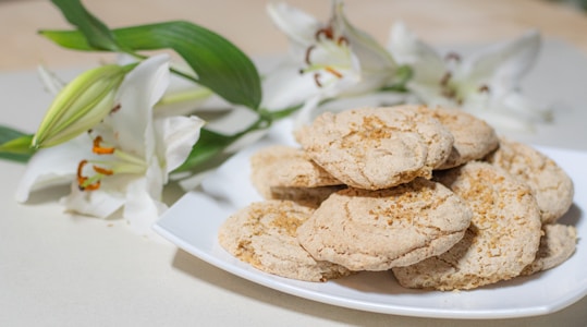 A white plate with a stack of homemade cookies dusted with brown sugar sits next to a bouquet of fresh, white lilies with green leaves. The setting is clean and simple, creating a contrast between the earthy tones of the cookies and the vibrant whites and greens of the flowers.