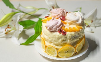 A layered cake is adorned with lemon slices around its base and topped with a variety of pastel-colored meringue swirls. There are also decorative peach slices and small red berry pieces on top. White lilies with green leaves are arranged beside the cake, enhancing the aesthetic presentation.