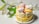 A layered cake is adorned with lemon slices around its base and topped with a variety of pastel-colored meringue swirls. There are also decorative peach slices and small red berry pieces on top. White lilies with green leaves are arranged beside the cake, enhancing the aesthetic presentation.