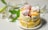 A layered cake is adorned with lemon slices around its base and topped with a variety of pastel-colored meringue swirls. There are also decorative peach slices and small red berry pieces on top. White lilies with green leaves are arranged beside the cake, enhancing the aesthetic presentation.