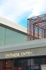 A modern building facade with a bilingual sign that reads 'ENTRADA ENTRY' in illuminated letters. The structure features glass panels and a geometric lattice design on the upper section. The sky is clear with a few clouds, adding to the architectural detail of the scene.