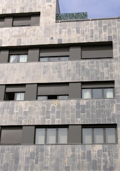 A section of a modern building facade with a stone tile exterior. The structure features multiple rectangular windows with dark frames and shutters. The surface textures of the stones are varied, with light and darker gray tones. A section of the building includes a green plant-covered balcony or rooftop.