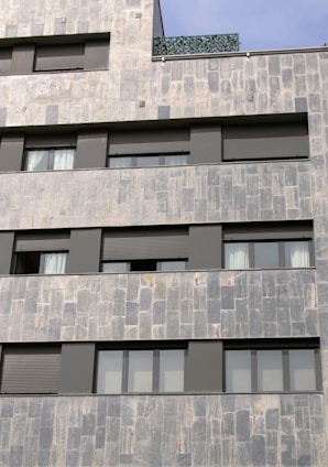 A section of a modern building facade with a stone tile exterior. The structure features multiple rectangular windows with dark frames and shutters. The surface textures of the stones are varied, with light and darker gray tones. A section of the building includes a green plant-covered balcony or rooftop.