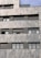 A section of a modern building facade with a stone tile exterior. The structure features multiple rectangular windows with dark frames and shutters. The surface textures of the stones are varied, with light and darker gray tones. A section of the building includes a green plant-covered balcony or rooftop.