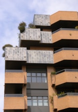A modern architectural building with a layered and geometric design featuring brick and concrete materials. The façade includes terrace spaces with greenery and plants, offering a blend of natural elements with urban architecture. Large windows add to the contemporary aesthetic.