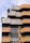 A modern architectural building with a layered and geometric design featuring brick and concrete materials. The façade includes terrace spaces with greenery and plants, offering a blend of natural elements with urban architecture. Large windows add to the contemporary aesthetic.