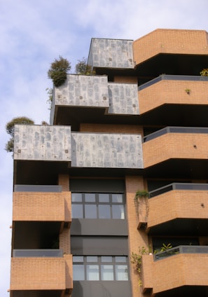 A modern architectural building with a layered and geometric design featuring brick and concrete materials. The façade includes terrace spaces with greenery and plants, offering a blend of natural elements with urban architecture. Large windows add to the contemporary aesthetic.