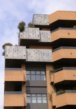 A modern architectural building with a layered and geometric design featuring brick and concrete materials. The façade includes terrace spaces with greenery and plants, offering a blend of natural elements with urban architecture. Large windows add to the contemporary aesthetic.