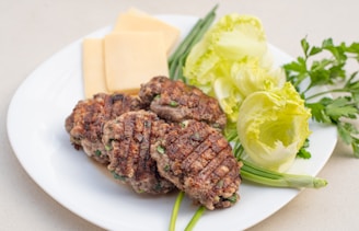Grilled patties with visible grill marks are arranged on a white plate alongside sliced cheese. Fresh green vegetables, including lettuce and herbs, provide a colorful garnish.