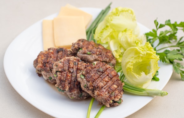 Grilled patties with visible grill marks are arranged on a white plate alongside sliced cheese. Fresh green vegetables, including lettuce and herbs, provide a colorful garnish.