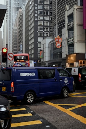 A busy urban street scene featuring tall buildings with commercial signage, a blue Brinks security van in the foreground crossing the intersection, and a red double-decker bus in the background. The scene is bustling with vehicles, and there are signs in different languages visible on the buildings.