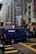 A busy urban street scene featuring tall buildings with commercial signage, a blue Brinks security van in the foreground crossing the intersection, and a red double-decker bus in the background. The scene is bustling with vehicles, and there are signs in different languages visible on the buildings.