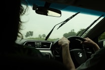 Inside a car, a person is driving while it rains outside. The windshield wipers are active, and the interior of the car shows a dashboard with air vents and a steering wheel displaying a Suzuki logo. Another individual, partially visible, is seated in the passenger seat.