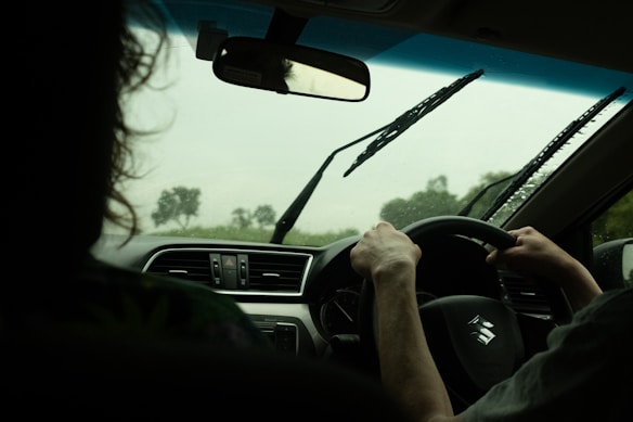 Inside a car, a person is driving while it rains outside. The windshield wipers are active, and the interior of the car shows a dashboard with air vents and a steering wheel displaying a Suzuki logo. Another individual, partially visible, is seated in the passenger seat.