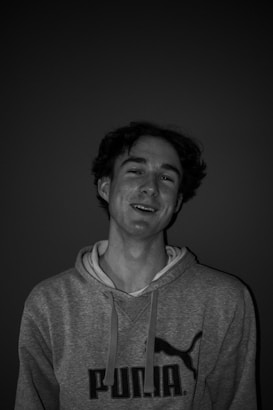 A person with wavy hair wearing a hoodie with a logo, smiling against a dark background. The lighting highlights the facial features, creating a contrast with the background.