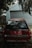 An old red Mitsubishi Libero GLXi car is parked in front of a closed garage door. The car appears to be used with some dents and scratches on the rear. It is surrounded by trees and leaves, indicating an outdoor environment. The light is dim, suggesting cloudy weather or shade.
