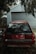 An old red Mitsubishi Libero GLXi car is parked in front of a closed garage door. The car appears to be used with some dents and scratches on the rear. It is surrounded by trees and leaves, indicating an outdoor environment. The light is dim, suggesting cloudy weather or shade.