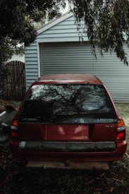 An old red Mitsubishi Libero GLXi car is parked in front of a closed garage door. The car appears to be used with some dents and scratches on the rear. It is surrounded by trees and leaves, indicating an outdoor environment. The light is dim, suggesting cloudy weather or shade.