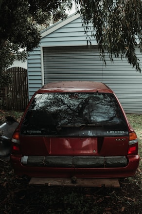 An old red Mitsubishi Libero GLXi car is parked in front of a closed garage door. The car appears to be used with some dents and scratches on the rear. It is surrounded by trees and leaves, indicating an outdoor environment. The light is dim, suggesting cloudy weather or shade.