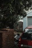 A red Mitsubishi car is parked beside a stack of bricks near a house. The house has light blue siding and a wooden gate. Dense foliage from trees surrounds the area, adding a sense of overgrowth and seclusion.