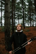 A young person stands in a forest, holding a camera tripod. The scene is set among tall trees with dark trunks and a forest floor covered in brown pine needles. The individual is wearing a dark jacket and has a content expression, suggesting a sense of tranquility or satisfaction.