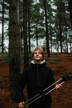 A young person stands in a forest, holding a camera tripod. The scene is set among tall trees with dark trunks and a forest floor covered in brown pine needles. The individual is wearing a dark jacket and has a content expression, suggesting a sense of tranquility or satisfaction.