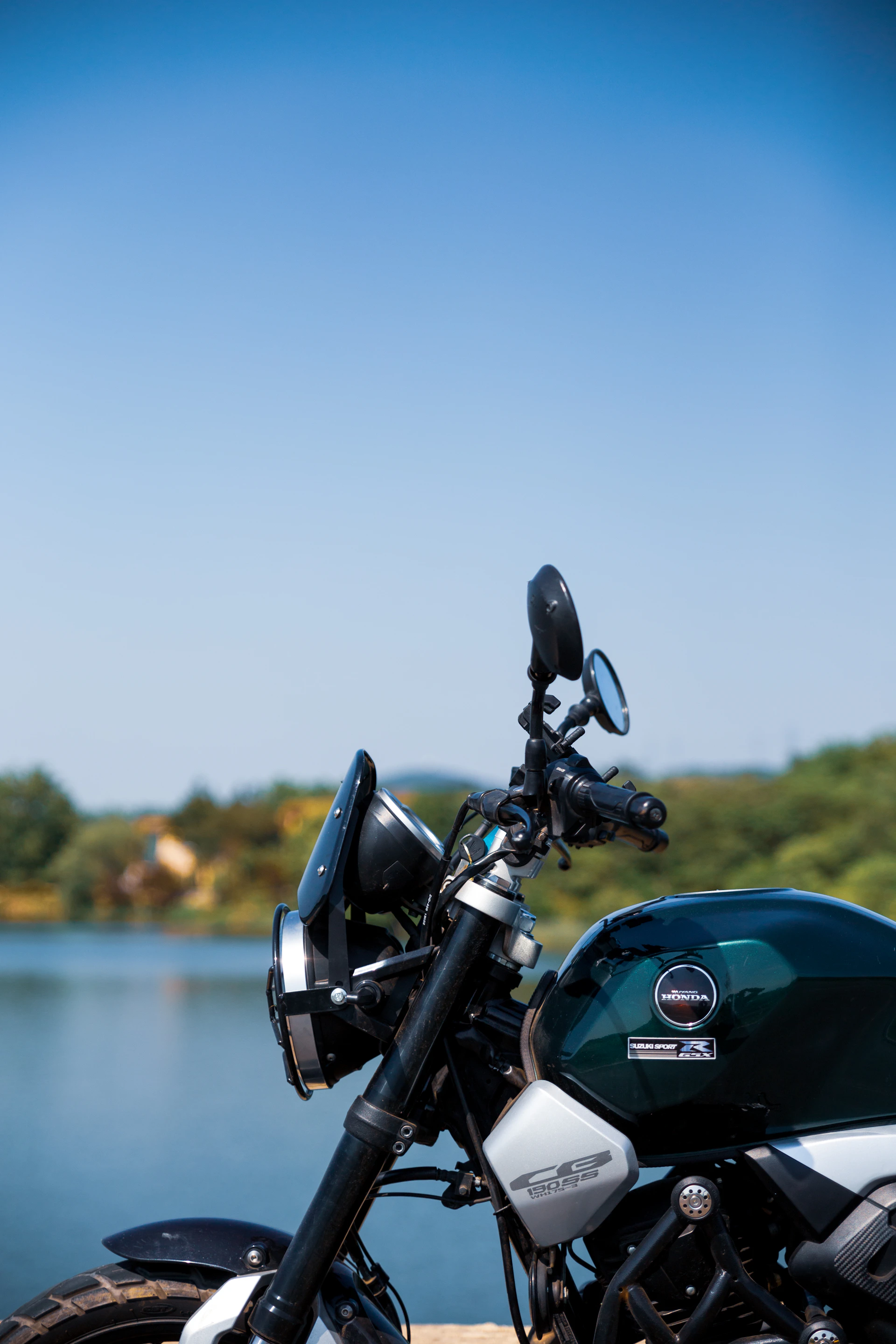 Close-up of a sleek motorcycle parked beside a crystal-clear lake, reflecting the surrounding pine trees and blue sky.