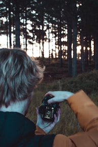 A person in an orange jacket is taking a photo with a DSLR camera in a forest. The background is filled with tall trees, and the sunlight peeks through, creating a serene and natural atmosphere.