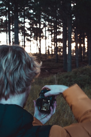 A person in an orange jacket is taking a photo with a DSLR camera in a forest. The background is filled with tall trees, and the sunlight peeks through, creating a serene and natural atmosphere.