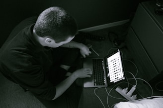 A person sitting on the floor using a laptop with images displayed on the screen. The setting appears to be a home or office environment with a carpeted floor, a wooden drawer nearby, and cables connected to a power strip.