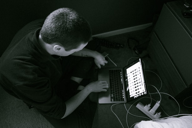A person sitting on the floor using a laptop with images displayed on the screen. The setting appears to be a home or office environment with a carpeted floor, a wooden drawer nearby, and cables connected to a power strip.