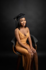 A person wearing a black graduation cap and a gold gown, sitting on a chair against a dark backdrop. The individual is smiling slightly and has their hands resting on their lap.