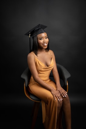A person wearing a black graduation cap and a gold gown, sitting on a chair against a dark backdrop. The individual is smiling slightly and has their hands resting on their lap.