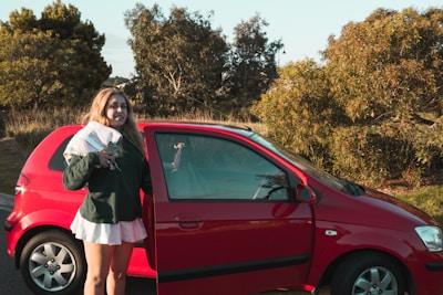 A person is standing beside an open red car door, holding keys and a folded towel. The car is parked on a road with greenery and trees in the background under a clear sky.