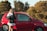 A person is standing beside an open red car door, holding keys and a folded towel. The car is parked on a road with greenery and trees in the background under a clear sky.