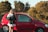 A person is standing beside an open red car door, holding keys and a folded towel. The car is parked on a road with greenery and trees in the background under a clear sky.