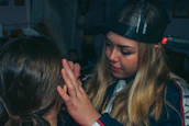 A person with long blonde hair is applying makeup or performing some form of facial grooming on another person. The setting appears cozy and informal, with dim lighting. The person working wears a black cap and is focused on the task.