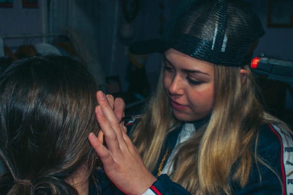 A person with long blonde hair is applying makeup or performing some form of facial grooming on another person. The setting appears cozy and informal, with dim lighting. The person working wears a black cap and is focused on the task.