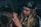 A person with long blonde hair is applying makeup or performing some form of facial grooming on another person. The setting appears cozy and informal, with dim lighting. The person working wears a black cap and is focused on the task.
