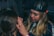 A person with long blonde hair is applying makeup or performing some form of facial grooming on another person. The setting appears cozy and informal, with dim lighting. The person working wears a black cap and is focused on the task.