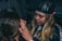 A person with long blonde hair is applying makeup or performing some form of facial grooming on another person. The setting appears cozy and informal, with dim lighting. The person working wears a black cap and is focused on the task.