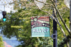 A street sign hangs from a post, featuring a stylized illustration of buildings and a sun with rays. The words 'Hilltop Business District' are prominently displayed in bold lettering. Surrounding the sign are out-of-focus tree branches with green leaves, and in the background, a traffic light shows a green signal.