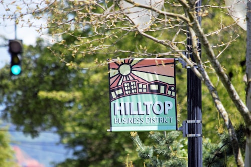 A street sign hangs from a post, featuring a stylized illustration of buildings and a sun with rays. The words 'Hilltop Business District' are prominently displayed in bold lettering. Surrounding the sign are out-of-focus tree branches with green leaves, and in the background, a traffic light shows a green signal.