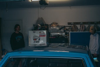 A garage scene with two individuals standing near a parked blue car. On the wall, there are coils of rope and various tools hanging. A white fridge with stickers is also visible, along with an assortment of other garage equipment and storage boxes.