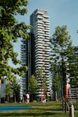 A modern high-rise residential building with unique staggered balconies is set amidst lush green trees. People can be seen enjoying the grassy park area in the foreground, engaging in activities like walking and resting. The scene is framed by clear blue skies and bright sunlight.
