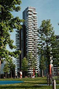 A modern high-rise residential building with unique staggered balconies is set amidst lush green trees. People can be seen enjoying the grassy park area in the foreground, engaging in activities like walking and resting. The scene is framed by clear blue skies and bright sunlight.