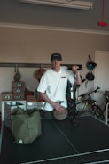A person wearing a white shirt and a cap is standing in a room with various items. The person is holding a basketball and seems to be next to an air pump. In the background, there are bicycles and shelves with boxes. A green bag is placed on a black table, which appears to be a table tennis table.