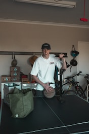 A person wearing a white shirt and a cap is standing in a room with various items. The person is holding a basketball and seems to be next to an air pump. In the background, there are bicycles and shelves with boxes. A green bag is placed on a black table, which appears to be a table tennis table.