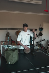 A person wearing a white shirt and a cap is standing in a room with various items. The person is holding a basketball and seems to be next to an air pump. In the background, there are bicycles and shelves with boxes. A green bag is placed on a black table, which appears to be a table tennis table.