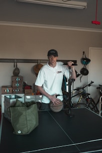 A person wearing a white shirt and a cap is standing in a room with various items. The person is holding a basketball and seems to be next to an air pump. In the background, there are bicycles and shelves with boxes. A green bag is placed on a black table, which appears to be a table tennis table.